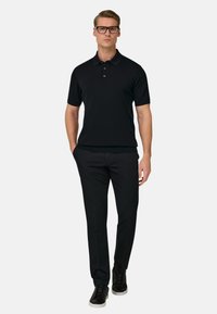 Homme portant un polo noir, un pantalon slim noir, des baskets noires à semelles blanches et des lunettes noires, debout avec une main dans la poche.