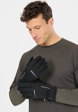 Mies, joka pukeutuu tummaan pitkähihaiseen paitaan ja laittaa mustia Endurance-käsineitä, joissa on heijastavat raidat, yksinkertaista valkoista taustaa vasten.