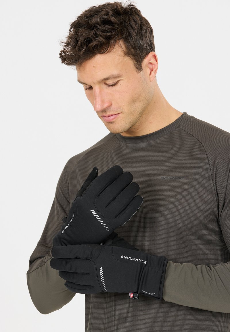 Mand iført mørk langærmet skjorte tager sorte Endurance-handsker med reflekterende striber på mod en ensartet hvid baggrund på.