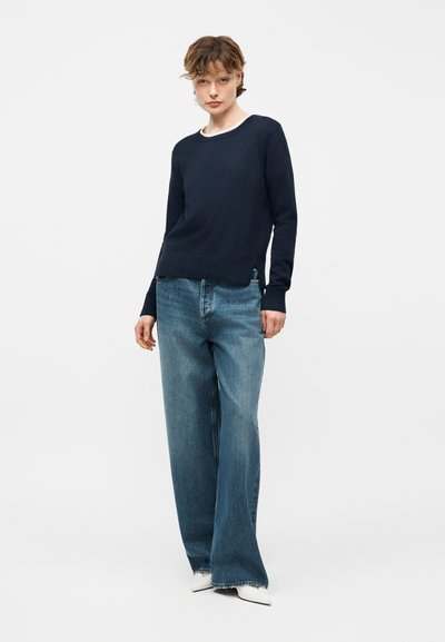 Jeune personne debout, portant un pull à manches longues bleu marine, un jean bleu ample et des chaussures blanches pointues, sur un fond blanc uni.