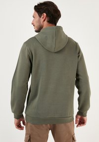 Olíva zöld kapucnis pulóver sima textúrával, bordázott mandzsettákkal és alsó széllel. Kanguru zseb és egyszerű design jellemzi.