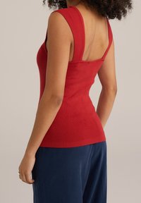 Czerwona, prążkowana bluzka na ramiączkach o szerokich paskach i dopasowanym kroju, z dekoltem w serek i teksturowanym wykończeniem. W połączeniu z granatowymi spodniami.