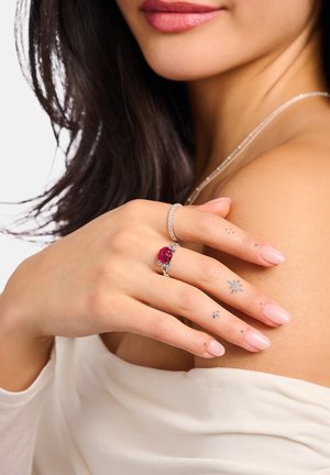 Frau berührt Schulter mit der Hand, die silberne Ringe trägt, darunter einen mit einem roten Rosendesign, und kleine Fingertattoos sind sichtbar.