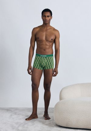 Groene boxershorts met een geometrisch patroon, voorzien van een zwarte tailleband met het "LACOSTE"-logo. Model staat blootsvoets op een licht tapijt.
