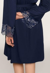 Granatowy szlafrok z długimi rękawami i wyszukanym koronkowym wykończeniem. Tkanina jest gładka, a w talii znajduje się pasek, co zapewnia dopasowany krój.