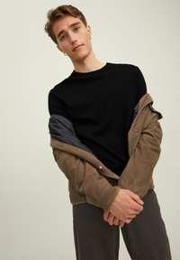 Pullover nero a collo rotondo indossato sotto una giacca in pelle scamosciata marrone, con un interno grigio. Il modello posa con le braccia incrociate, indossando pantaloni scuri.