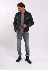 Zwarte leren jas met voorzakken, grijs T-shirt, lichtblauwe distressed jeans en zwarte enkellaarsjes. Model staat casual.