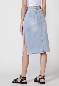 Denim krilo v svetlo modri barvi z razporkom spredaj, z obliko zadnjega žepa in ravno silhueto, dopolnjeno z črnimi sandali na vezalke.
