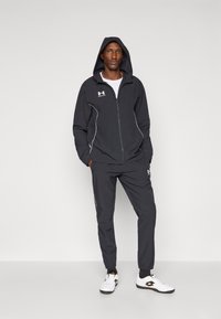 Compleu athletic negru cu glugă, cu accente albe și închidere cu fermoar. Modelul poartă pantofi albi cu margini negre și șosete.