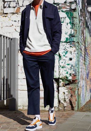 Chaqueta azul marino sobre sudadera blanca con cremallera y camiseta naranja. Pantalones azul marino y zapatillas con detalles en naranja y blanco. Fondo texturizado.