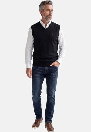Uomo con capelli grigi e barba, indossa una camicia bianca abbottonata, un maglione senza maniche nero, jeans scuri e scarpe di pelle marrone, in piedi e guarda verso destra.