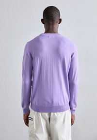 Lilafärgad stickad tröja med rund halsringning, ribbad nederkant och muddar. Slät textur, långa ärmar, bärs över ljusa shorts.