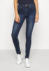 Jeans ajustados de un azul oscuro, hechos de denim con una textura suave, que cuentan con un bolsillo delantero, un botón de cobre y pasadores para cinturón, combinados con zapatillas blancas.