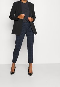 Černý blazer přes tmavé tričko, doplněný námořnickými kostkovanými úzkými kalhotami. Černé lodičky dokončují celkový outfit.