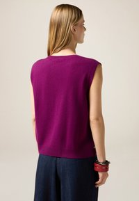 Bezrekawowy sweter zrobiony na drutach z żywej fioletowej wełny, z okrągłym dekoltem i lekko luźnym krojem, zestawiony z ciemnymi dżinsami.