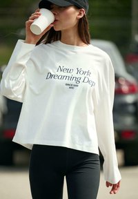 Fehér, hosszú ujjú ing "New York Dreaming Dept." felirattal, fekete léggingsekkel párosítva; a modell kávéscsészét tart, laza szabadtéri környezetben.