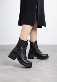 Botas de tobillo de cuero negro con tacón grueso, cordones en el frente y cremallera lateral. Presentan un material texturizado y acentos en contraste.