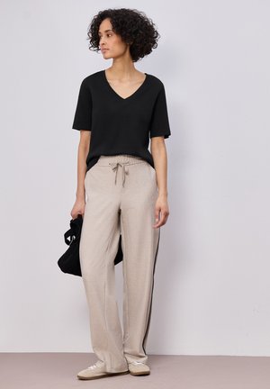 Kobieta z krótkimi kręconymi włosami, ubrana w czarną bluzkę z krótkim rękawem i dekoltem w serek, beżowe spodnie z sznurkiem i paskiem po boku, trzyma czarną torebkę, stoi na tle jednolitej ściany.