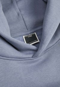 Hoodie gris avec une texture douce et une poche kangourou ; présente une étiquette noire avec le texte "JACK & JONES" sur le col.