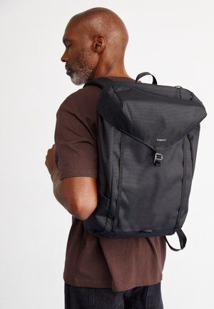 Black backpack with a textured finish, top flap closure, adjustable straps, and a front buckle. Worn on a person’s back.