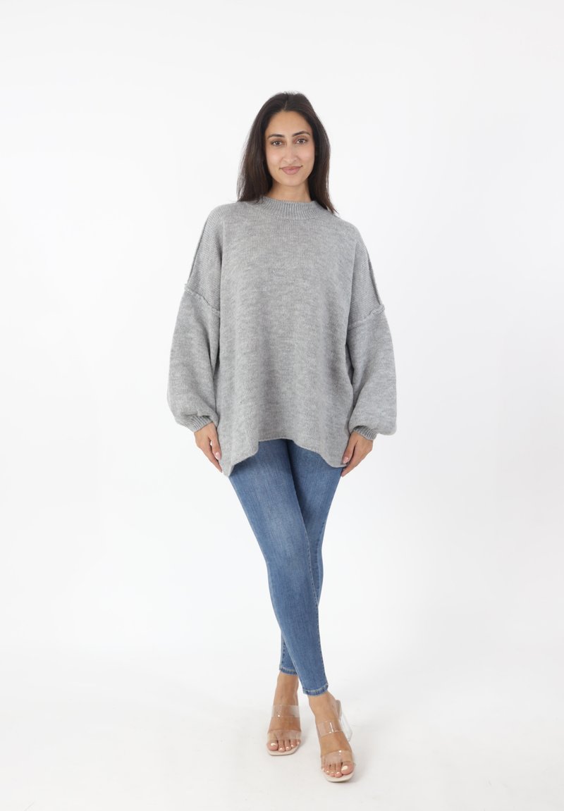 Maglione oversize grigio chiaro lavorato a maglia con maniche a palloncino, abbinato a jeans skinny blu e sandali con tacco trasparente su uno sfondo neutro.