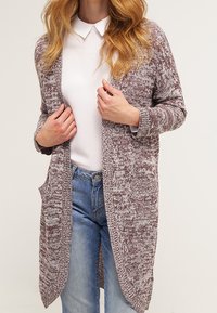 Donna che indossa un cardigan a maglia bicolore marrone e bianco sopra una camicia bianca con colletto e jeans azzurri chiari, mentre regola la parte anteriore del cardigan.