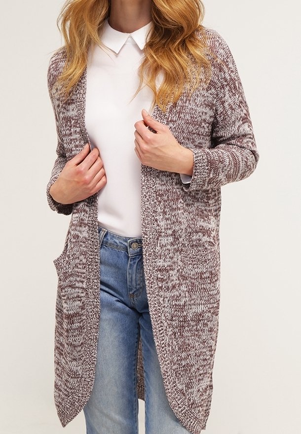 Donna che indossa un cardigan a maglia bicolore marrone e bianco sopra una camicia bianca con colletto e jeans azzurri chiari, mentre regola la parte anteriore del cardigan.