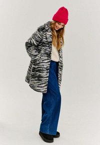 Kabát z vlnenej zmesi s čierno-bielym pruhovaným vzorom, kombinovaný s modrými širokými džínsami a jasne ružovou rebrovanou čiapkou. Čierne platformové topánky.