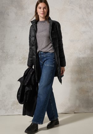 Manteau en doudoune matelassé noir, pull gris, jeans bleus et bottines noires. Manteau tenu dans la main gauche; le modèle se tient contre un fond texturé.