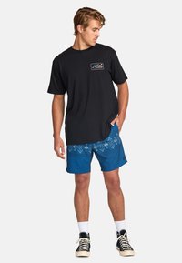 T-shirt en coton noir avec un graphique de montagne coloré, associé à un short bleu avec un imprimé géométrique. Le modèle porte des baskets noires et des chaussettes blanches.