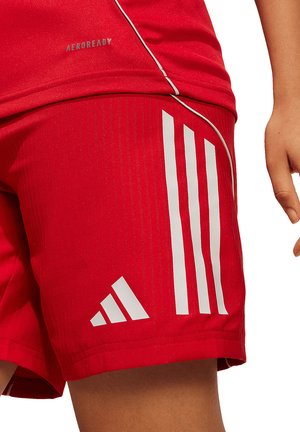 Zbliżenie osoby noszącej czerwone sportowe spodenki Adidas z trzema białymi paskami oraz część czerwonej koszulki z napisem Aeroready.