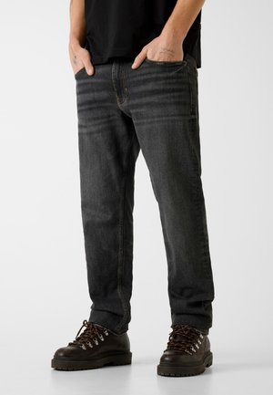 Unterkörper einer Person, die ausgewaschene schwarze gerade Jeans und braune Leder-Wanderschuhe trägt, Hände teilweise in den Taschen, vor weißem Hintergrund.