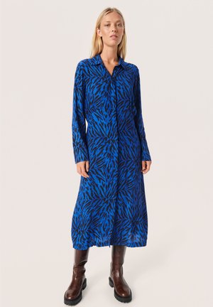 Blau- und schwarz gemustertes Hemdkleid mit langen Ärmeln, Kragen und Knopfleiste; strukturierten Stoff; kombiniert mit braunen Stiefeletten bis zum Knie.