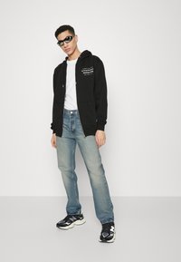 Sudadera negra con cremallera y texto blanco, combinada con una camiseta blanca, jeans azul claro y zapatillas negras. El modelo lleva gafas de sol oscuras.