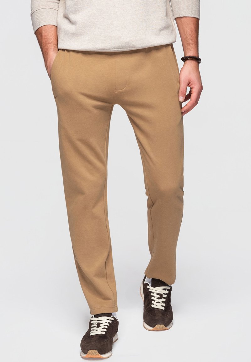 Uomo che indossa pantaloni casual beige, maglione chiaro, sneaker marroni, con le mani in tasca, in piedi contro uno sfondo chiaro e uniforme.