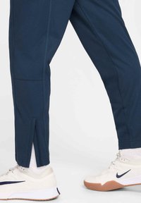 Tamsiai mėlynos sportinės kelnės siaurėjančiu siluetu, su plyšeliais kulkšnies srityje ir kontrastingomis siūl�ėmis, derinamos su baltais sportbačiais ir baltomis kojinėmis.