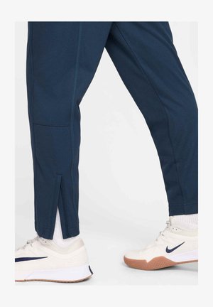 Navy blue athletic pants with a tapered design, featuring a side slit at the ankle and contrasting stitching. Worn with white sneakers.