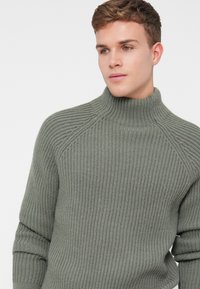 Ribota, zaļa augsta apkakles džemperis ar brīvu piegriezumu. Iezīmē augstu apkakli un raglanu piedurknēm, izgatavots no mīkstas, strukturētas auduma.