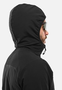 Homme portant une veste à capuche noire et des lunettes de soleil réfléchissantes, vu de profil sur un fond blanc uni.