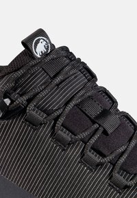 Nahaufnahme eines schwarzen Sneakers mit dünnen weißen Streifen, gepolstertem Zungenbereich, geschnürten Schnürsenkeln und einem kleinen Mammut-Logo auf dem Zuglasche.