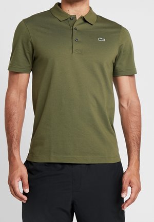 Muž nosící olivově zelené polo tričko s krátkým rukávem a malým logem krokodýla na levé straně hrudi a černé kalhoty.