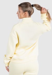 Gul sweatshirt med hög krage, oversized passform, ribbade ärmslut och mjuk textur; bärs med matchande underdelar; sedd bakifrån.