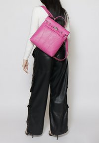 Roze leren rugzak met een gestructureerd ontwerp, voorzien van een handvat bovenop, verstelbare schouderbanden en metalen hardware accenten, geplaatst op een zwarte outfit.