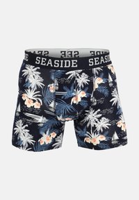 Marineblå boxershorts med hvide og ferskenfarvede blomster- og palmemønstre, sejlbåde og en sort talje med teksten "SEASIDE" i hvide bogstaver.