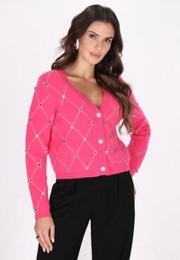 Cardigan fucsia con scollatura a V, caratterizzato da un motivo a rombi, decorato con strass colorati e chiusura a bottoni, indossato su pantaloni neri.