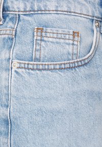 Blugi din denim albastru deschis, cu un buzunar frontal cu cusături decorative și detaliu cu buton argintiu, prezentând un finisaj moale și texturat.