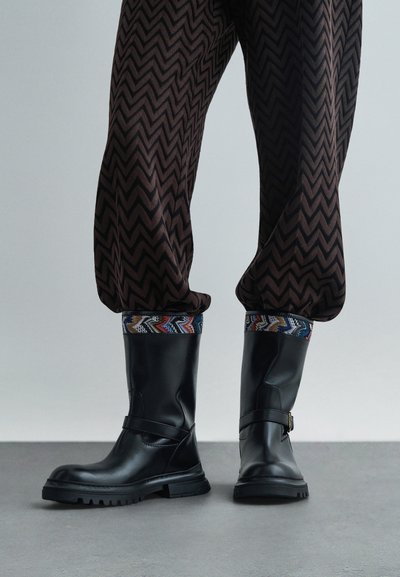 Black leather boots with a chunky sole, featuring a side strap and colorful patterned trim at the top. Worn with patterned dark pants.