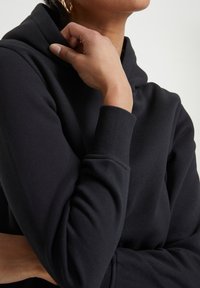Hoodie noir avec une texture douce, doté d'une capuche à cordon de serrage et de poignets côtelés. Le design présente une coupe décontractée et un style minimaliste.