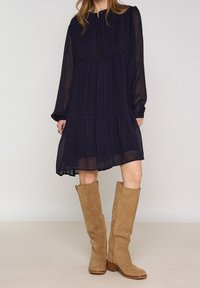 Marinblå klänning med långa genomskinliga ärmar, rund halsringning och volangkjol. Ett par knähöga stövlar i tanfärgad mocka med träklackar.