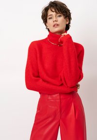 Roter gestrickter Rollkragenpullover mit lockerer Passform und langen Ärmeln. Kombiniert mit hochtaillierten roten Lederhosen, die eine glatte Textur aufweisen.